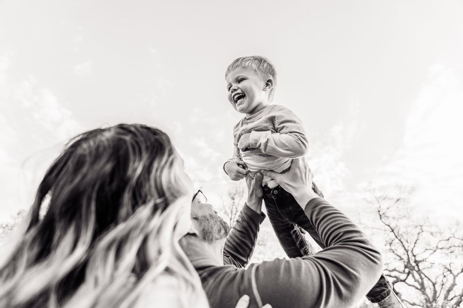 Black and white photo of a joyful toddler being lifted in the air by a woman outdoors.
