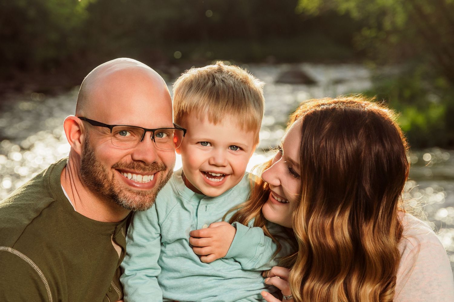 Happy family portrait with bald father, toddler boy, and brunette mother smiling outdoors by a sunlit river.