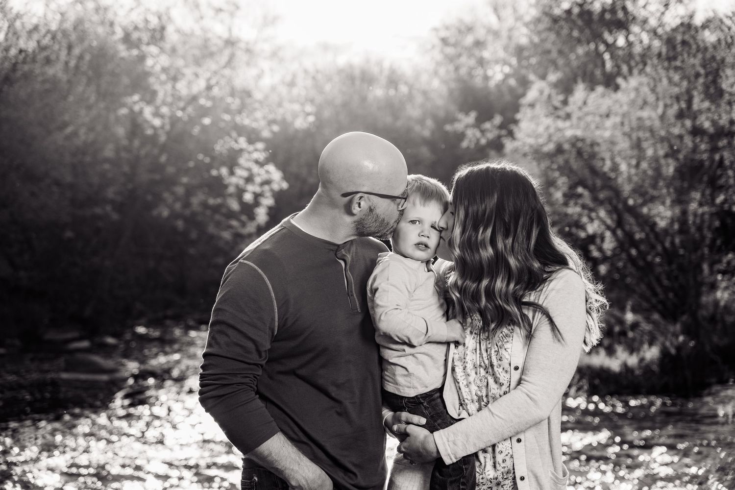 Black and white family portrait of parents kissing toddler outdoors near trees and stream.