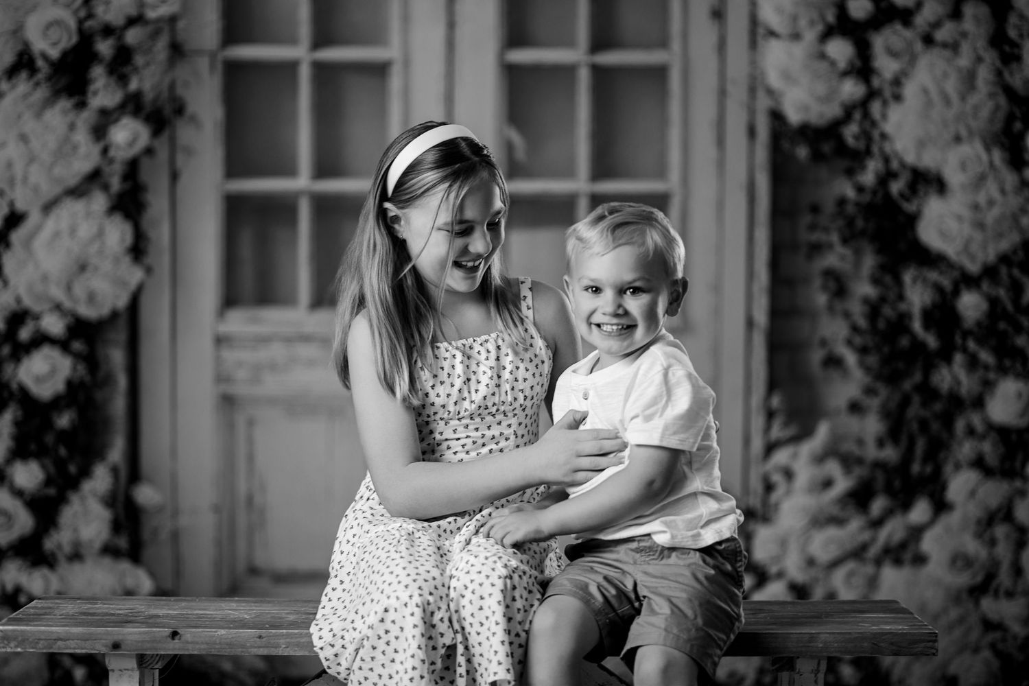 Black and white photo of a young girl laughing with her toddler brother seated on a bench before floral backdrop.