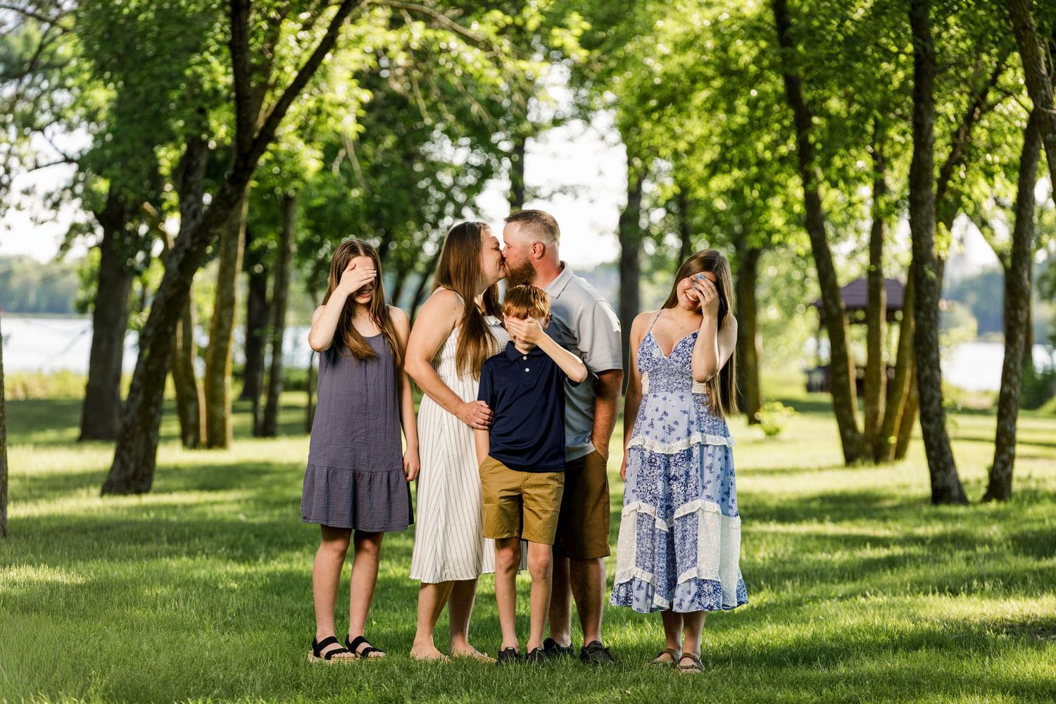 Family of five posing outdoors in a lush green park, parents kissing while children playfully cover their eyes.