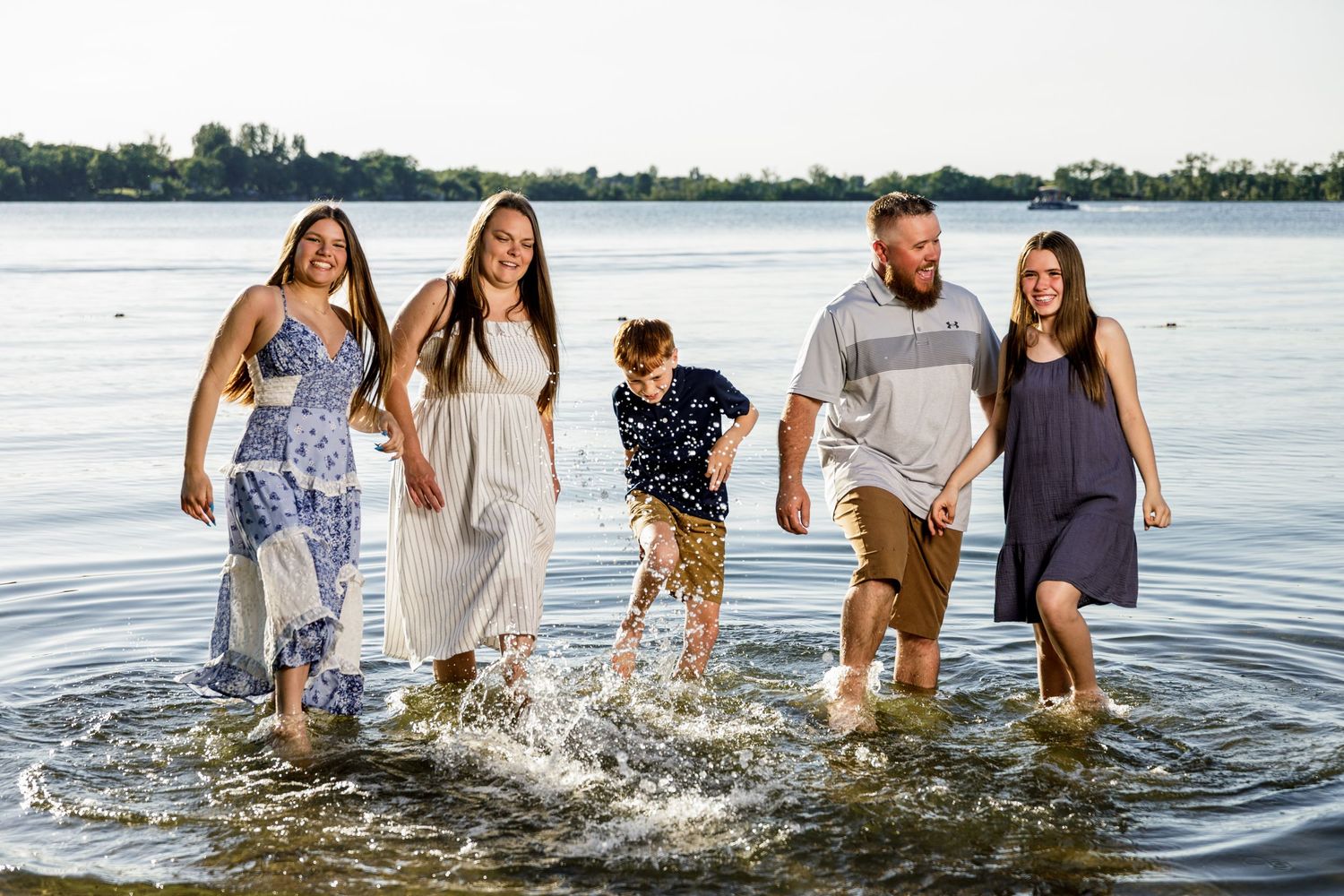 Family of five splashing in shallow lake water, smiling and holding hands on a sunny day.