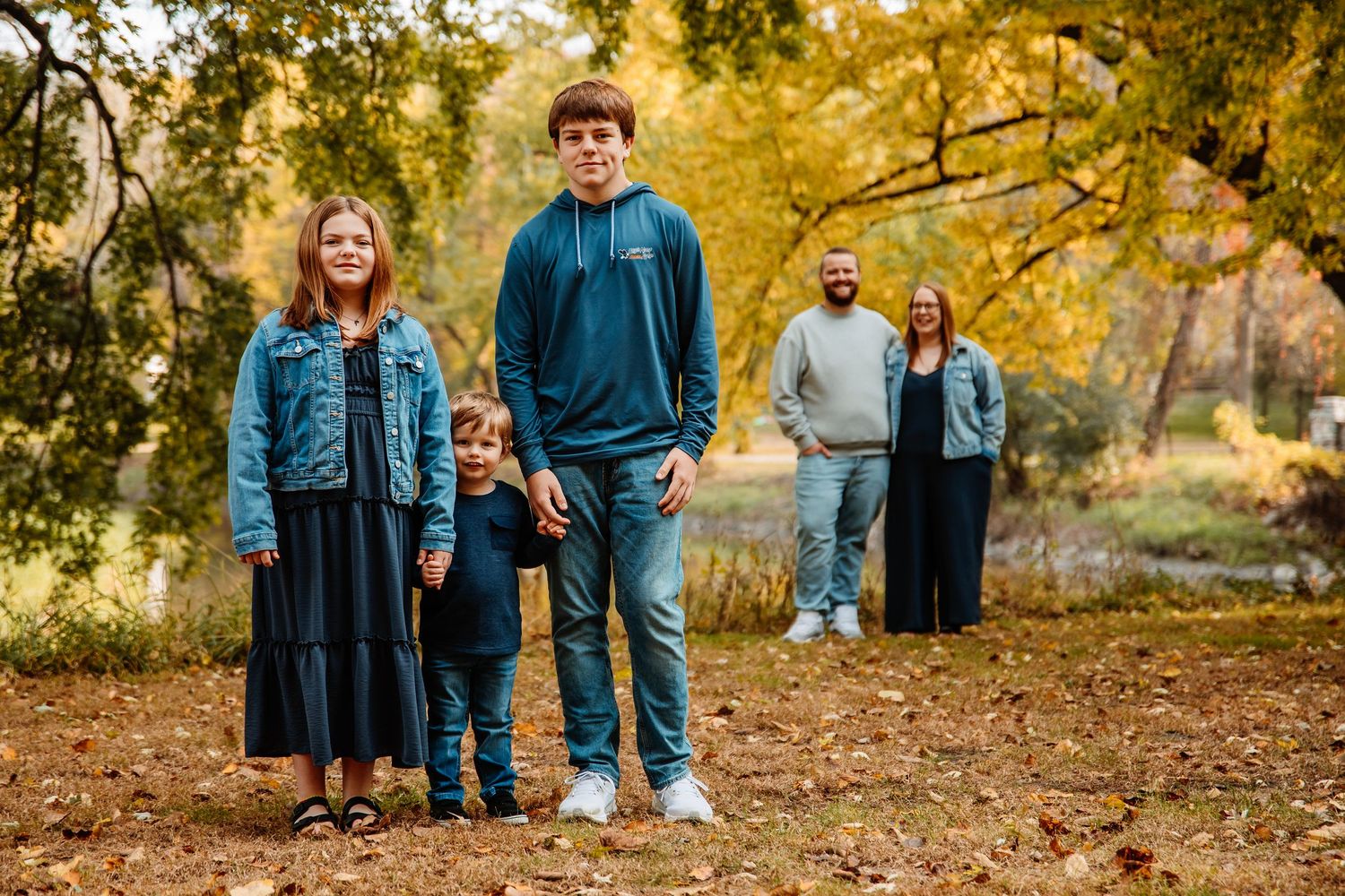 Family wearing coordinated blue outfits poses together during golden autumn photoshoot in park.