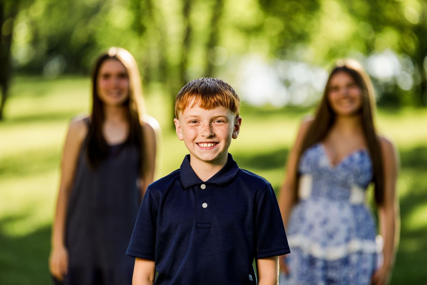 Smiling redhead boy in navy polo shirt stands in front of two teenage girls in a sunny green park.