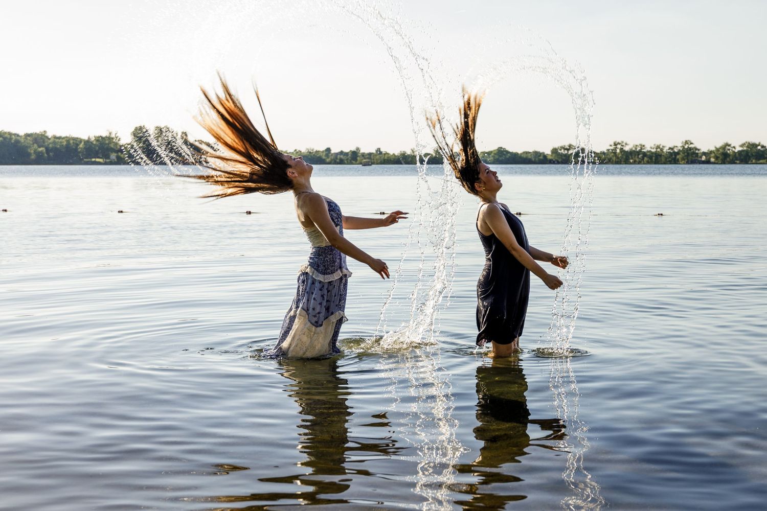 Two girls flipping wet hair creating water arcs while standing in a lake at sunset.