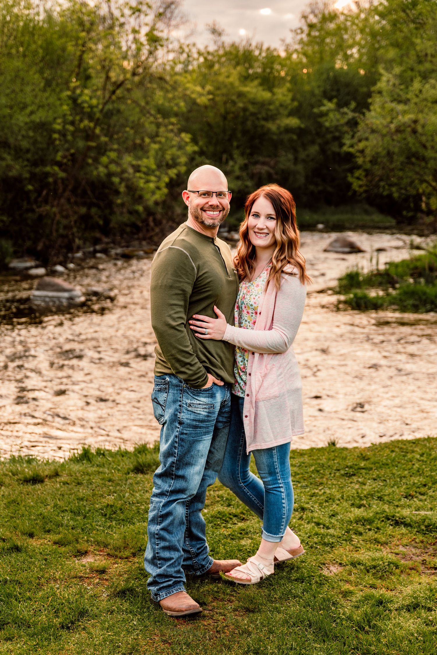 A smiling couple poses together near a peaceful creek surrounded by lush green trees during golden hour.