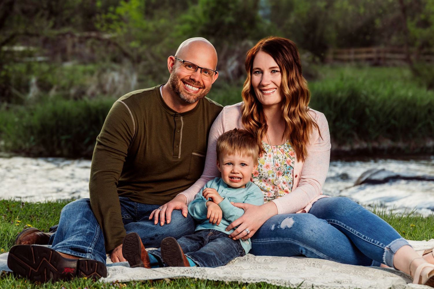 Happy young family with baby sitting on blanket in green park for outdoor portrait photo session.