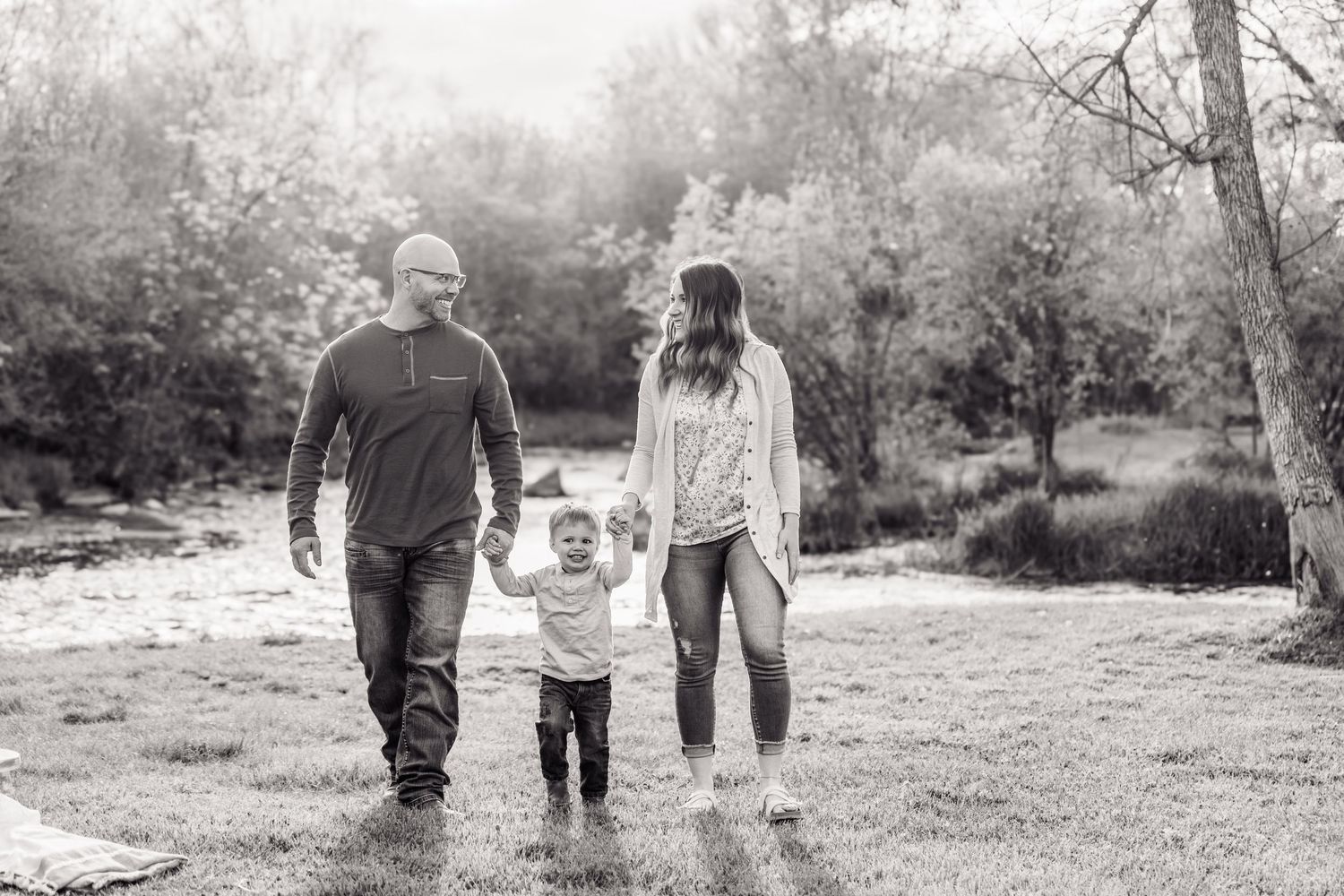 Black and white photo of a family of three walking in a park, parents holding toddler's hands.