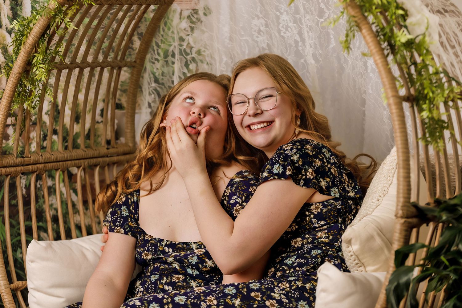 Two smiling girls in matching floral dresses hugging in a wicker egg chair with greenery backdrop.