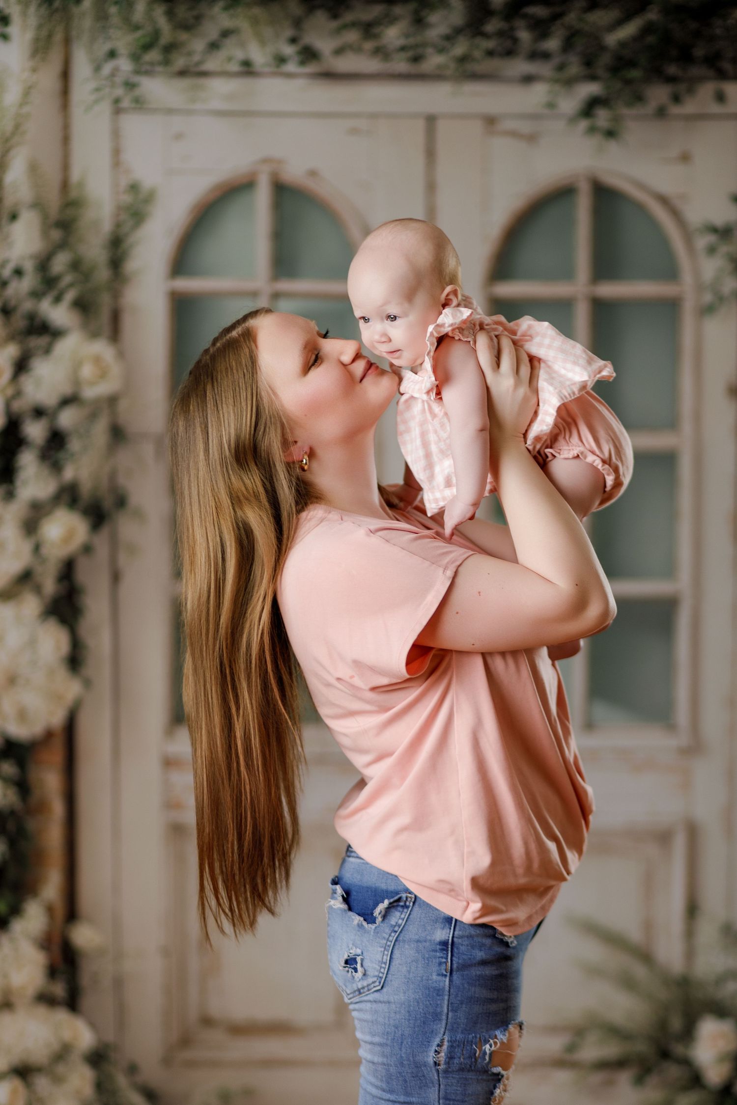 A loving mother lifts her baby girl in a pink dress for a kiss in a floral studio setting.