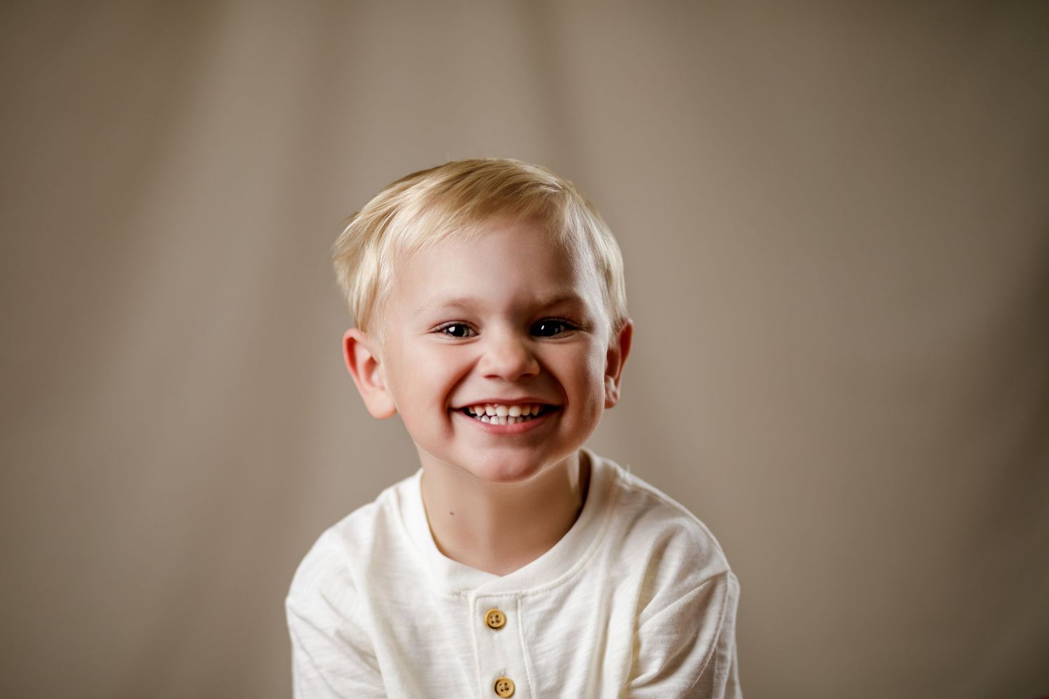 Adorable smiling blonde toddler boy in a white henley shirt posing for a cheerful portrait photo.