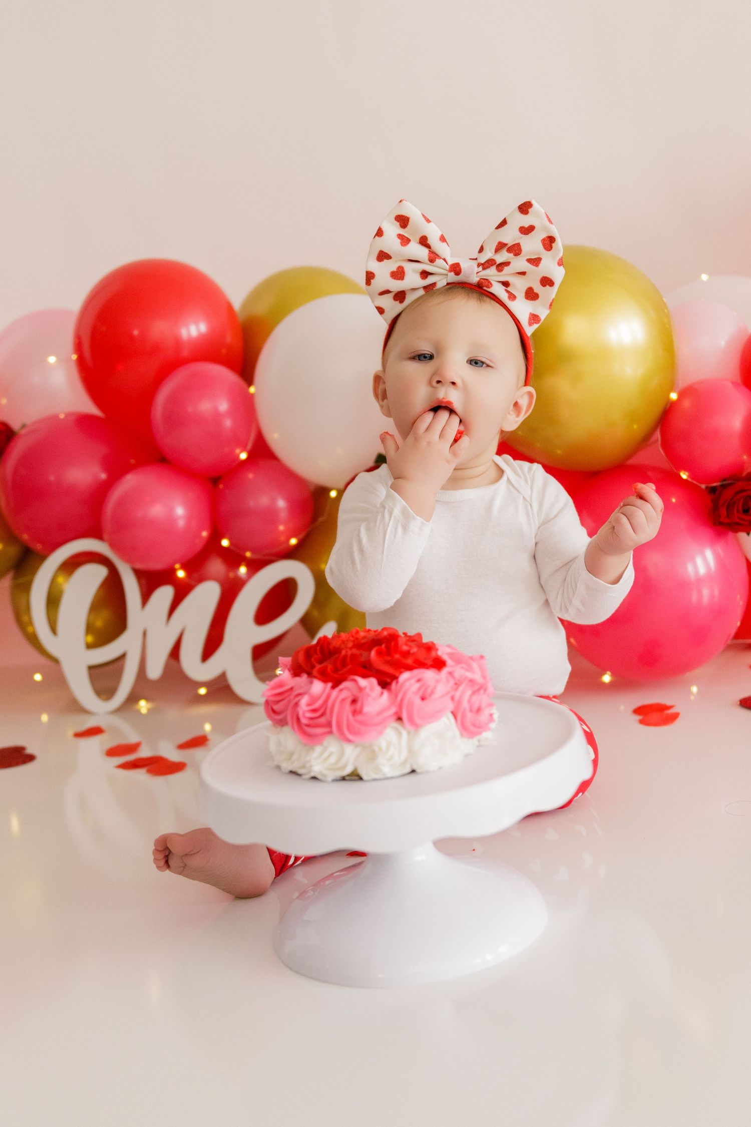 Baby in white dress on cake smash photo shoot with red and gold balloon backdrop.
