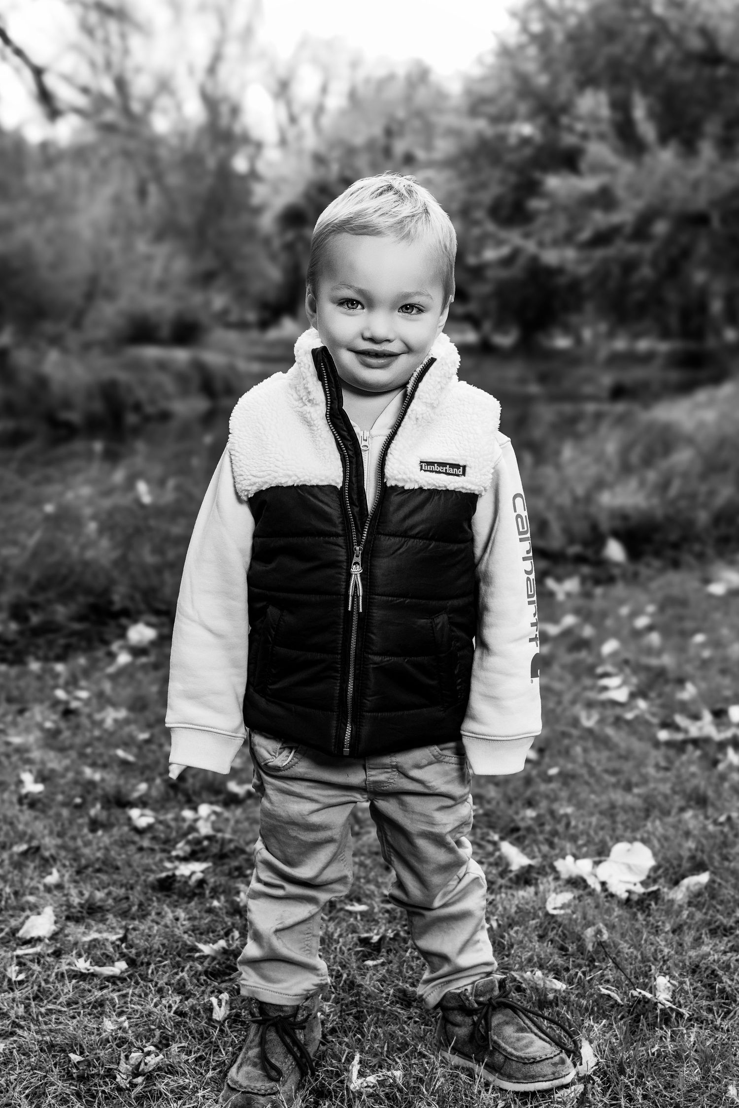 Young child in black vest and light pants smiling while standing outdoors in autumn.