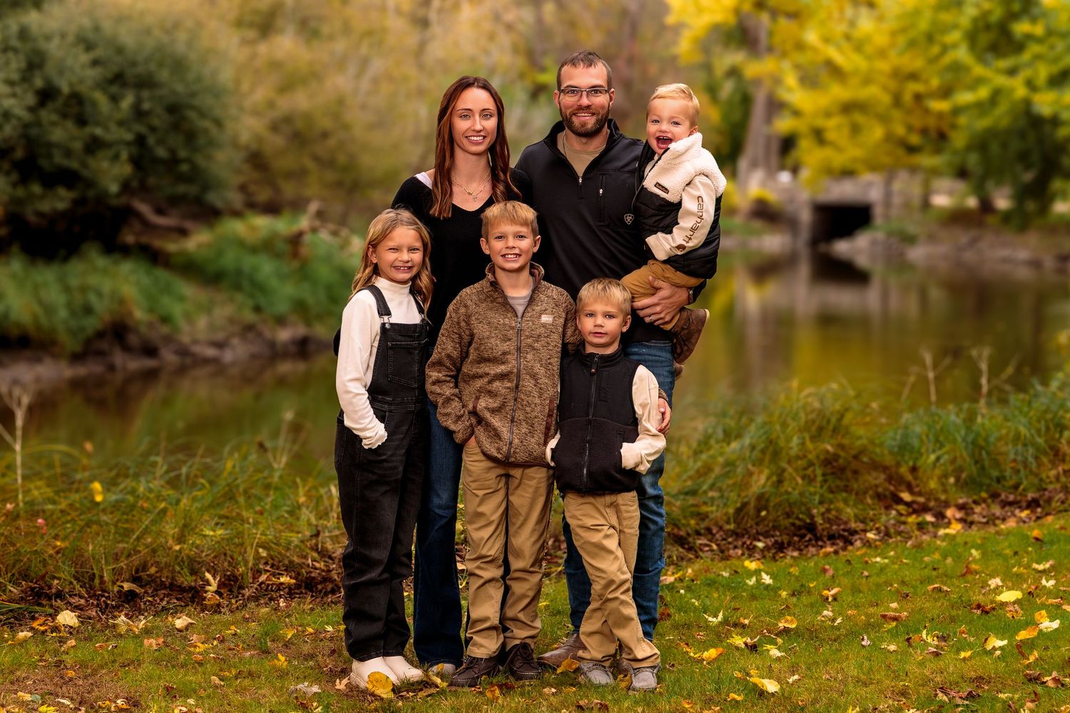 A family portrait taken in autumn by a pond with fall foliage in the background.
