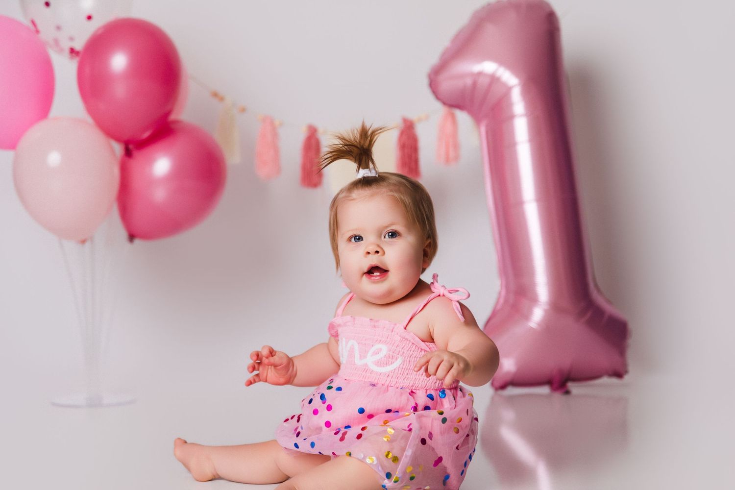 Baby in pink outfit surrounded by festive pink balloons and number one decoration.