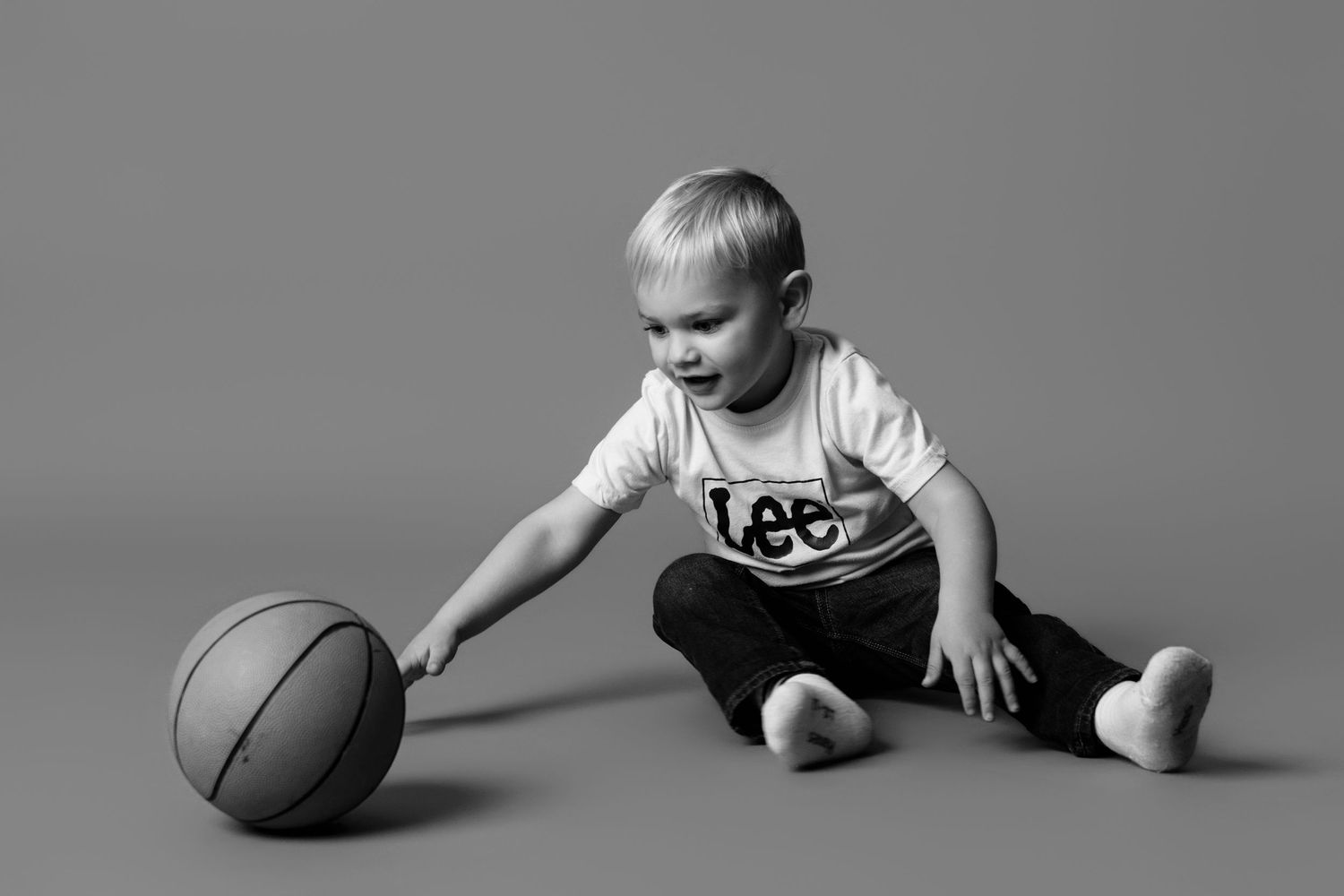A child sits on the floor with a basketball in a minimalist studio setting.