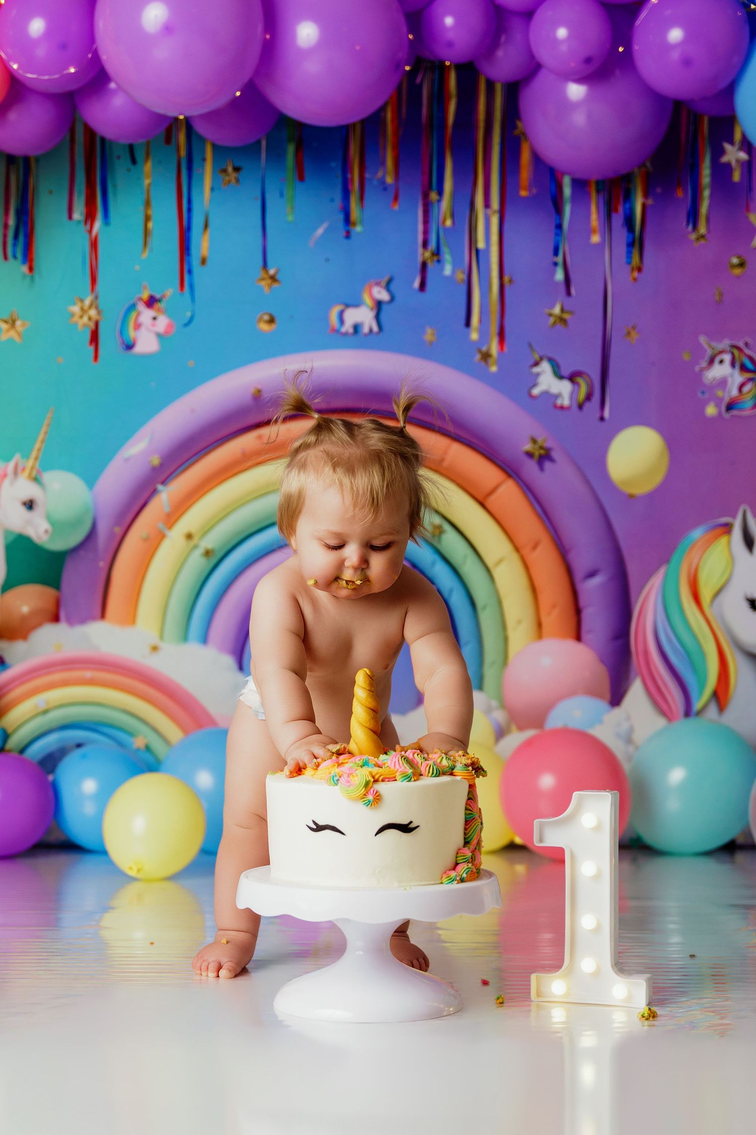 Baby girl smashing unicorn birthday cake at colorful first birthday party with rainbow and balloon decorations.