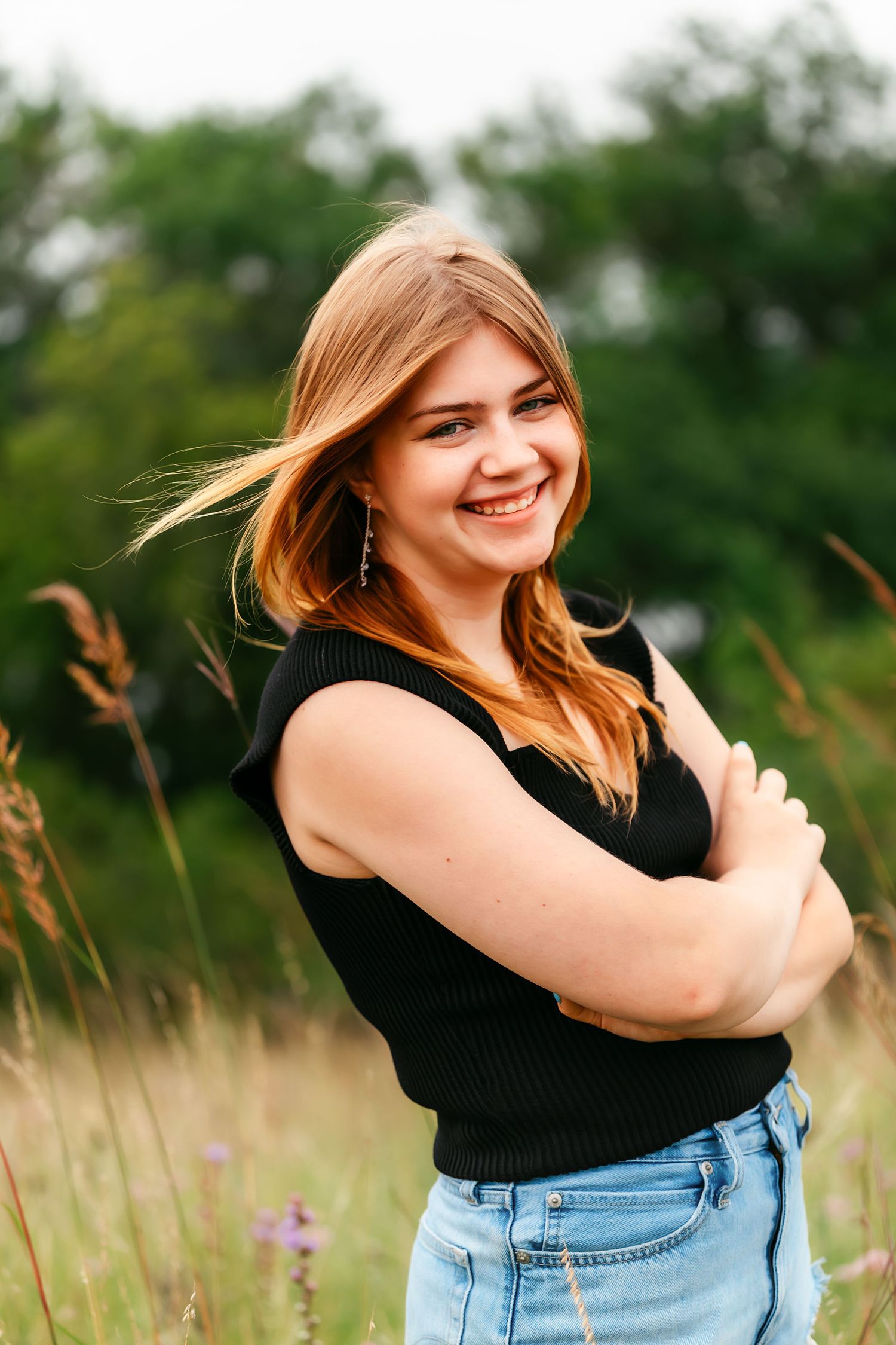Woman in black sleeveless top and denim poses joyfully in a field of tall grass on a summer day.