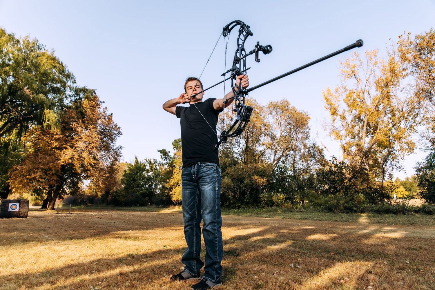 Archer in casual attire draws compound bow while practicing outdoors during autumn with golden trees in background.