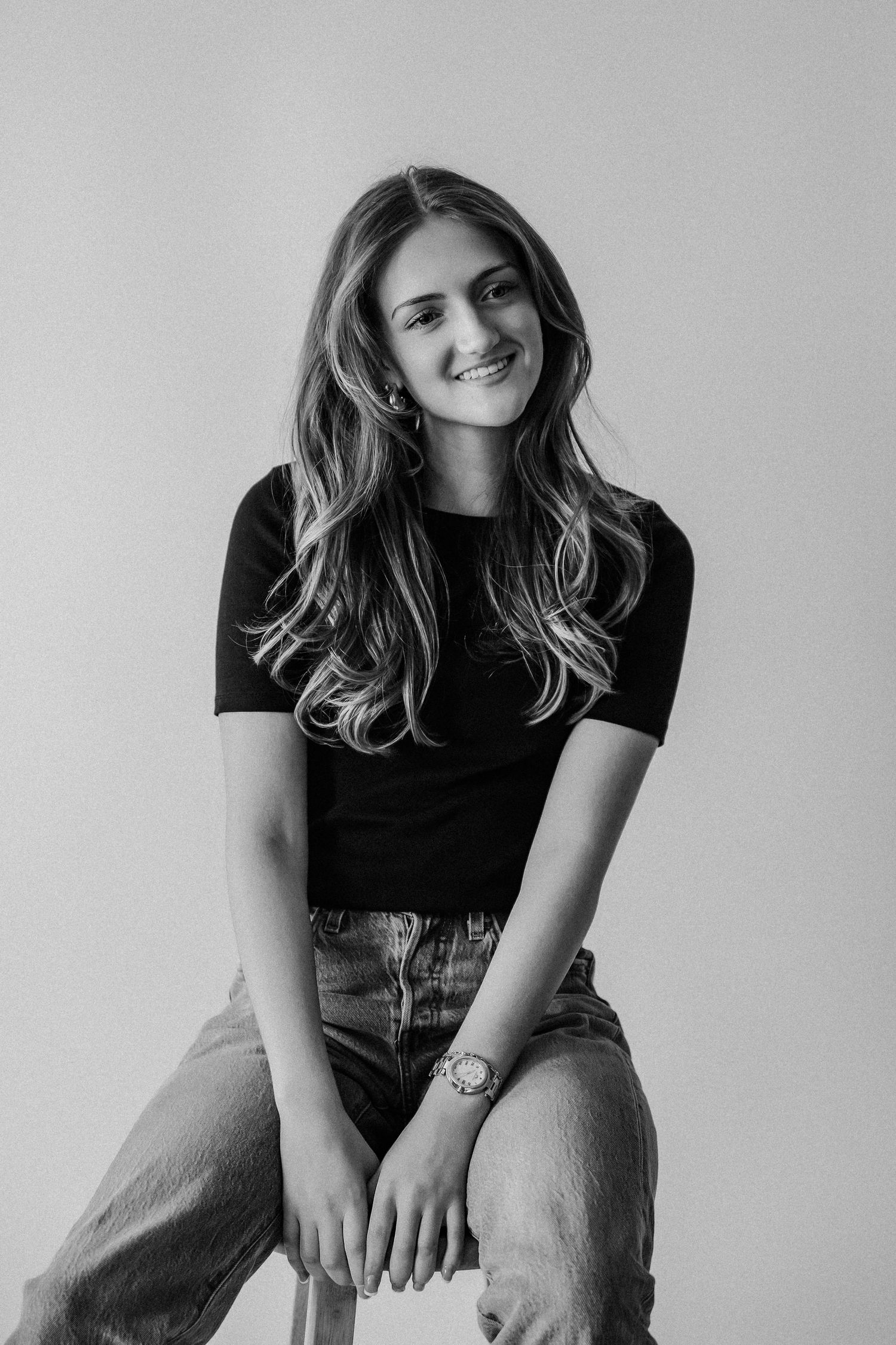 Black and white studio portrait of a smiling young woman in black t-shirt and jeans seated on a stool.