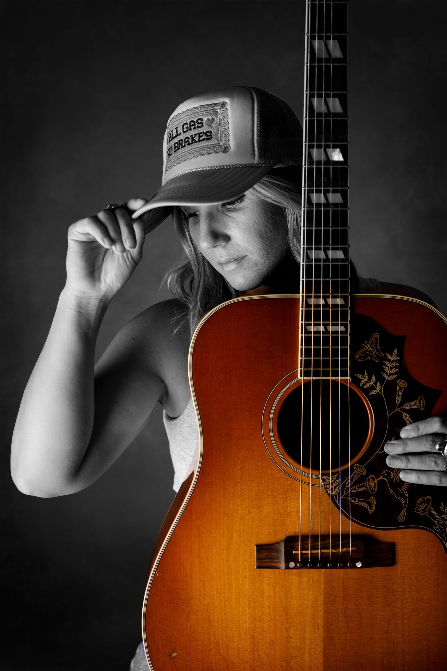 Female guitarist tips trucker cap beside vintage sunburst acoustic guitar in dramatic black and white portrait.