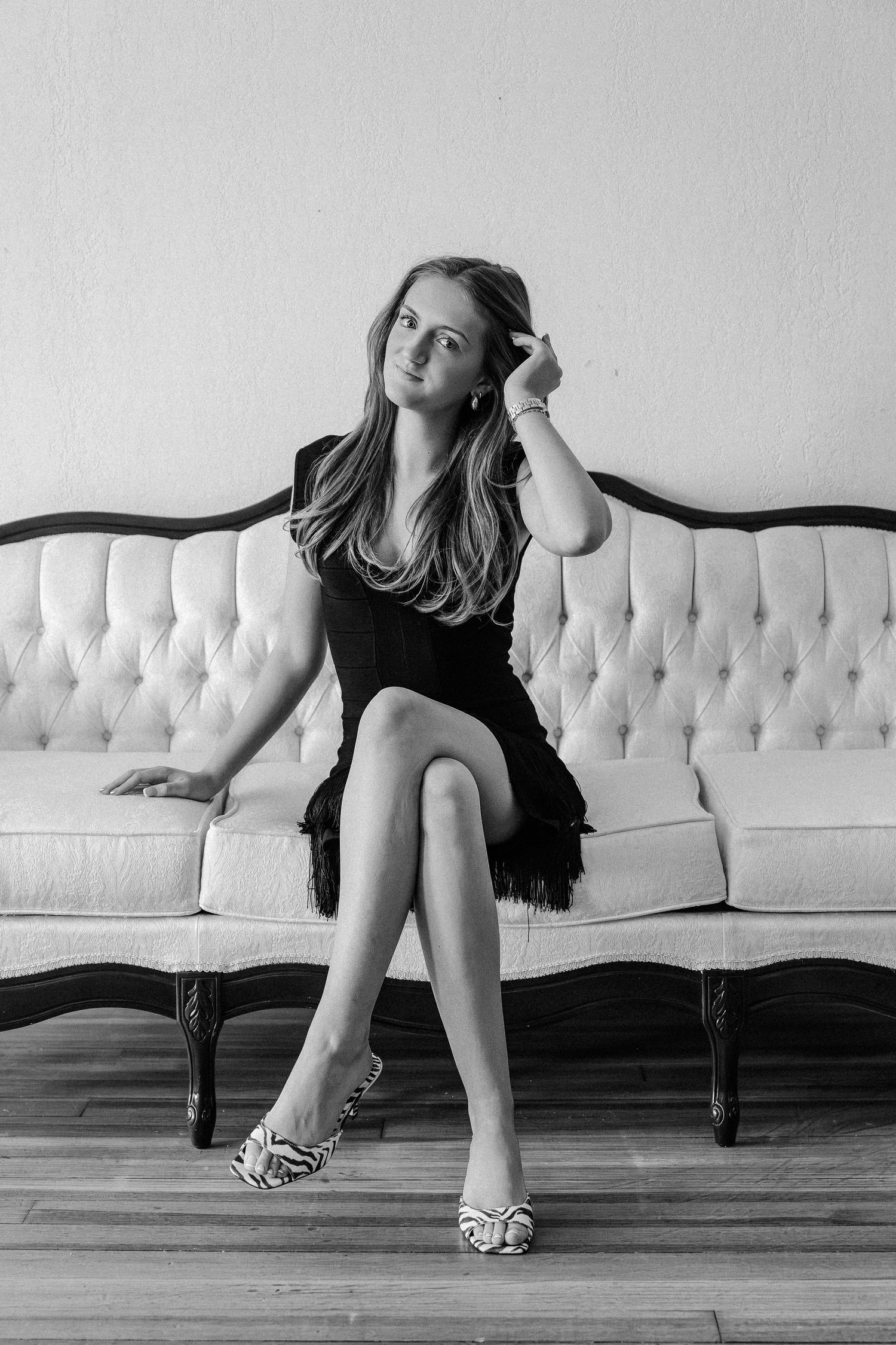 Black and white portrait of a young woman in black fringe dress seated on a tufted sofa with zebra heels.