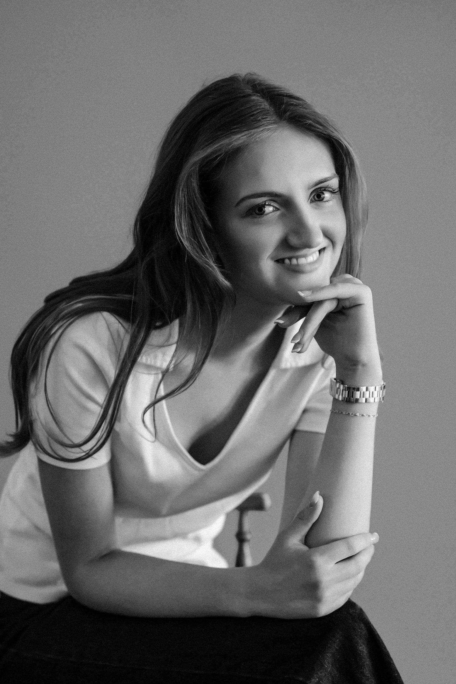 Black and white portrait of a smiling young woman resting chin on hand wearing a white top and bracelet.