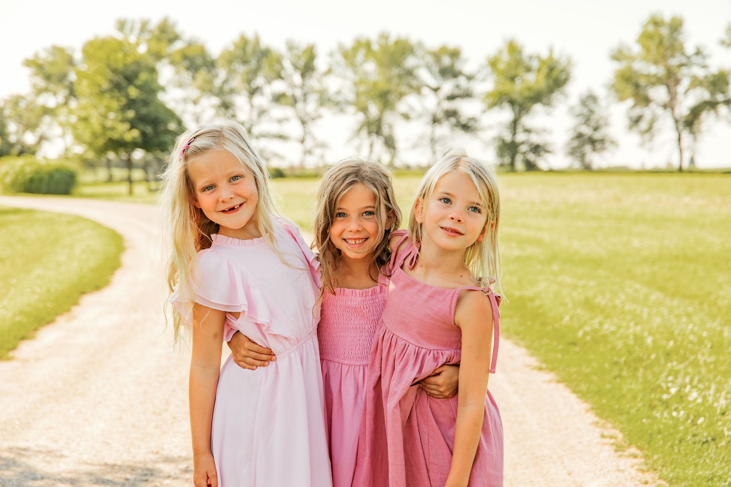 Three friends in pink dresses posing together outdoors on a sunny summer day.