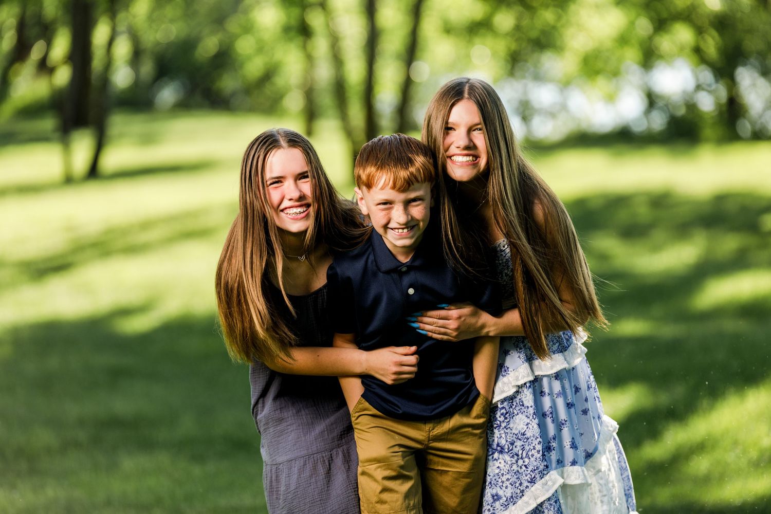 Two sisters hugging their younger brother outdoors in a lush green park during a family photo session.