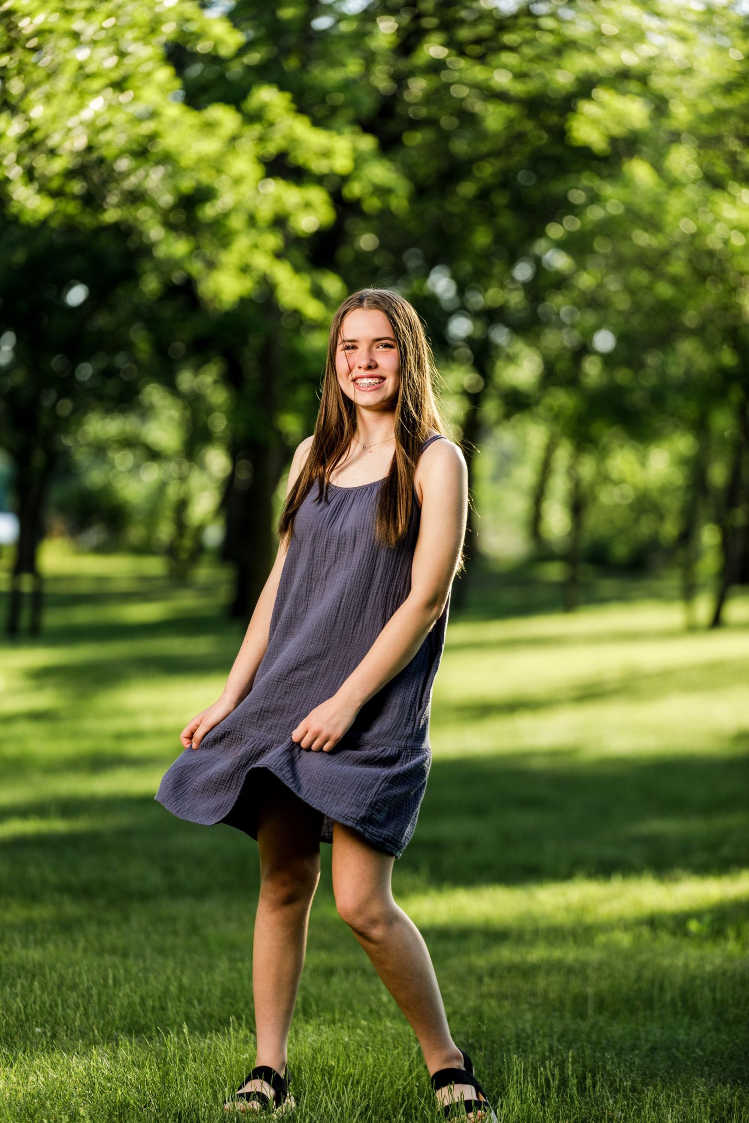 Smiling teenage girl in a gray dress standing on green grass in a sunlit park.