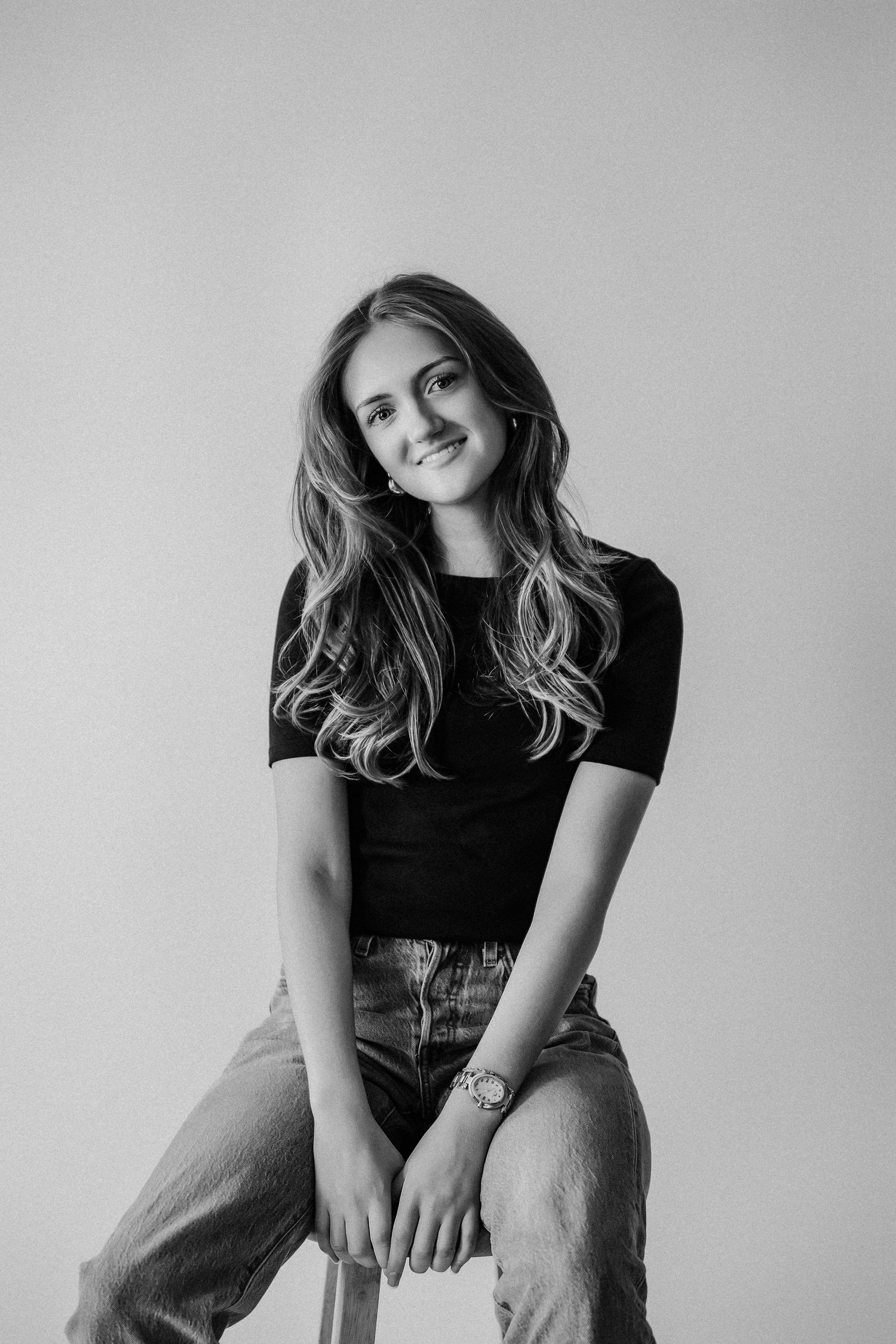 Black and white photo of a young woman in black top and jeans sitting on a stool, smiling softly.