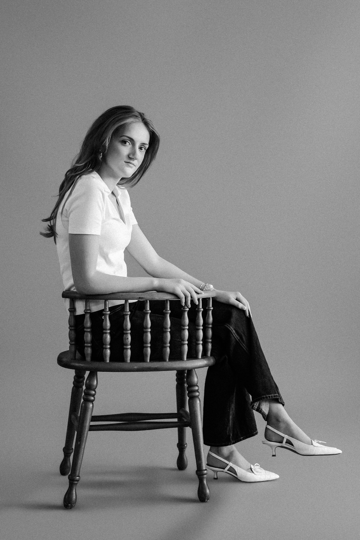 Black and white portrait of young woman in white blouse and dark pants seated on vintage wooden chair.