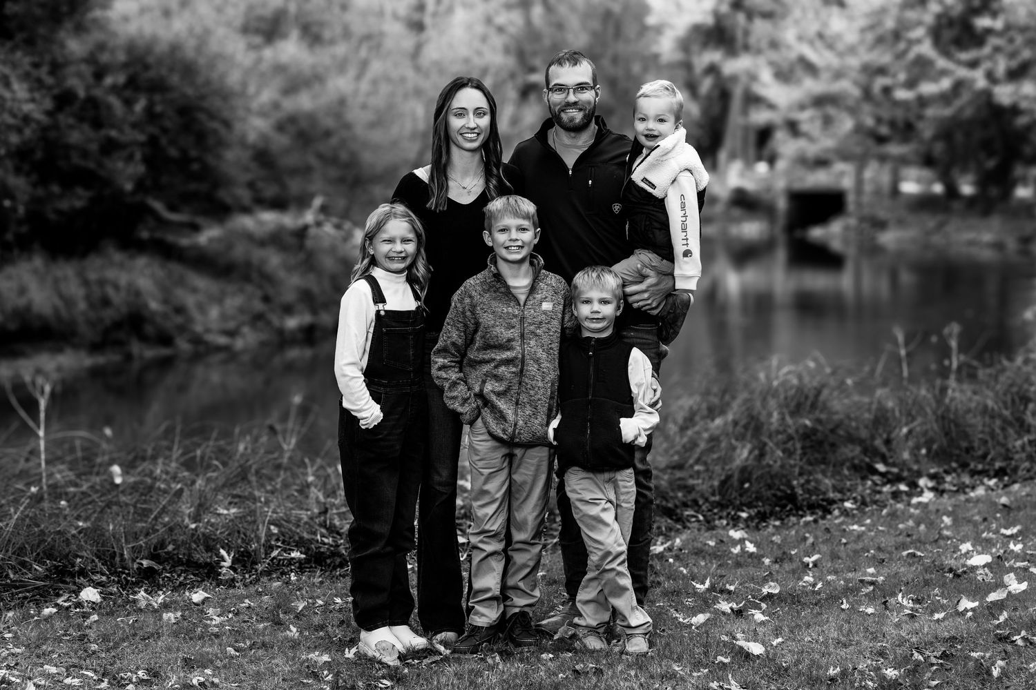Family group poses together outdoors during fall season with trees and fallen leaves in background.