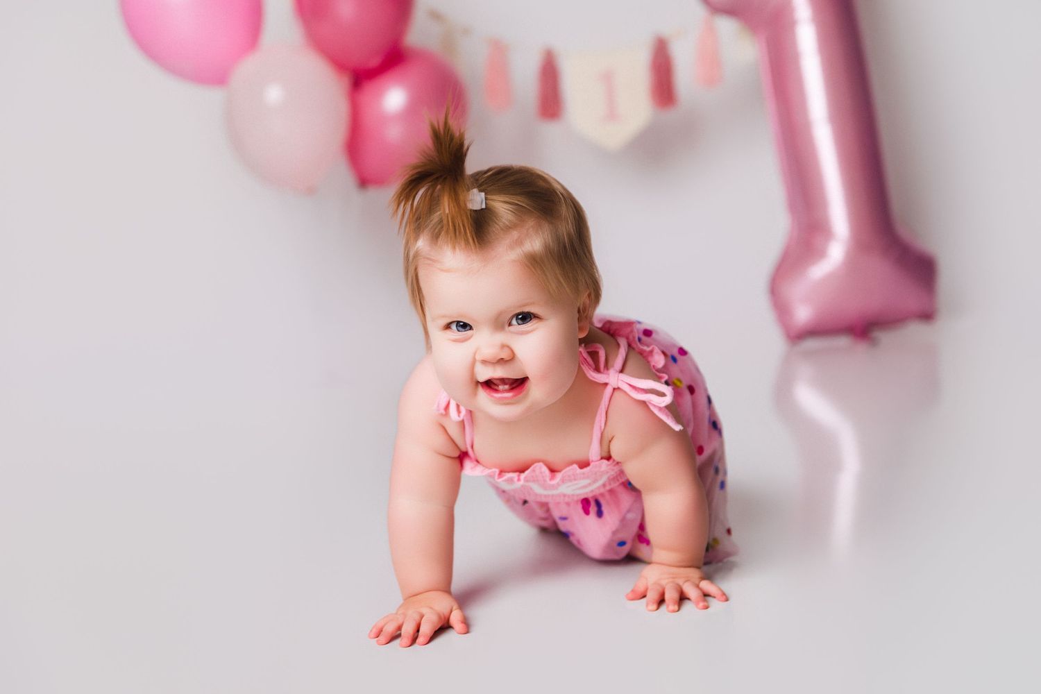 Baby in pink outfit crawling near pink and white balloon decorations.