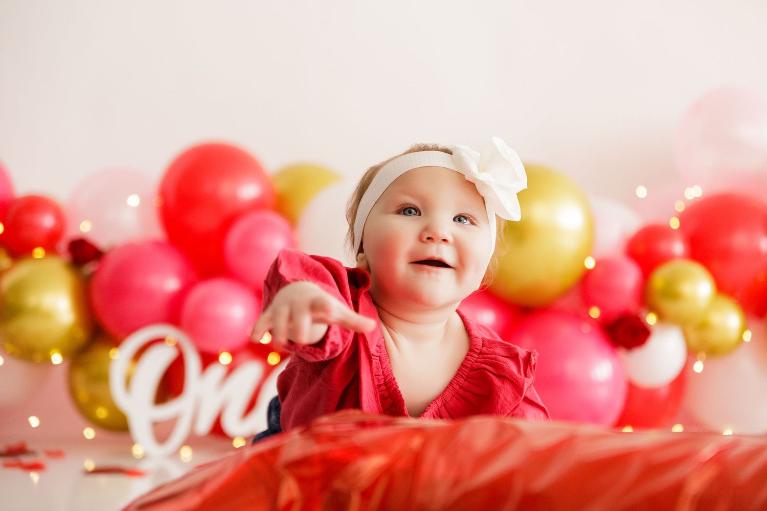 Baby in red outfit sits among pink and gold balloons during Valentine's Day photo session.