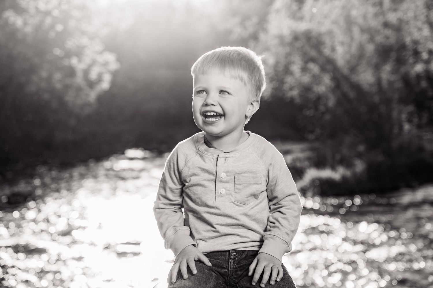 Black and white portrait of a laughing toddler boy sitting outdoors near a sunlit river surrounded by trees.