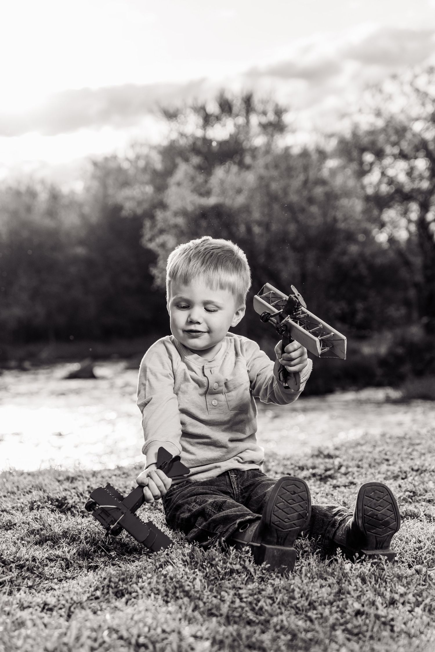 Black and white photo of a young boy sitting on grass, playing with wooden toy airplanes outdoors.