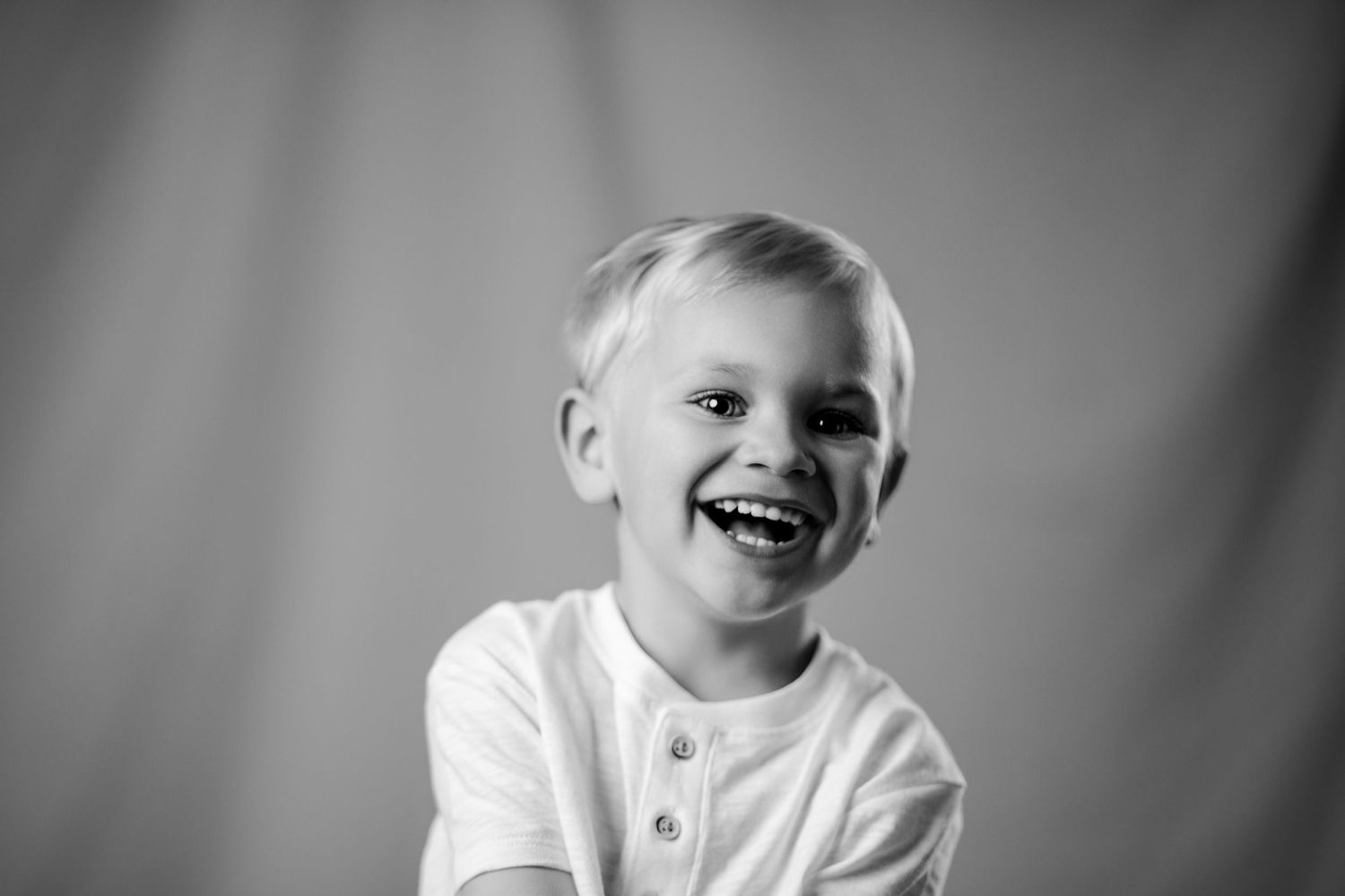 Adorable laughing blonde boy in white shirt captured in a black and white studio portrait.