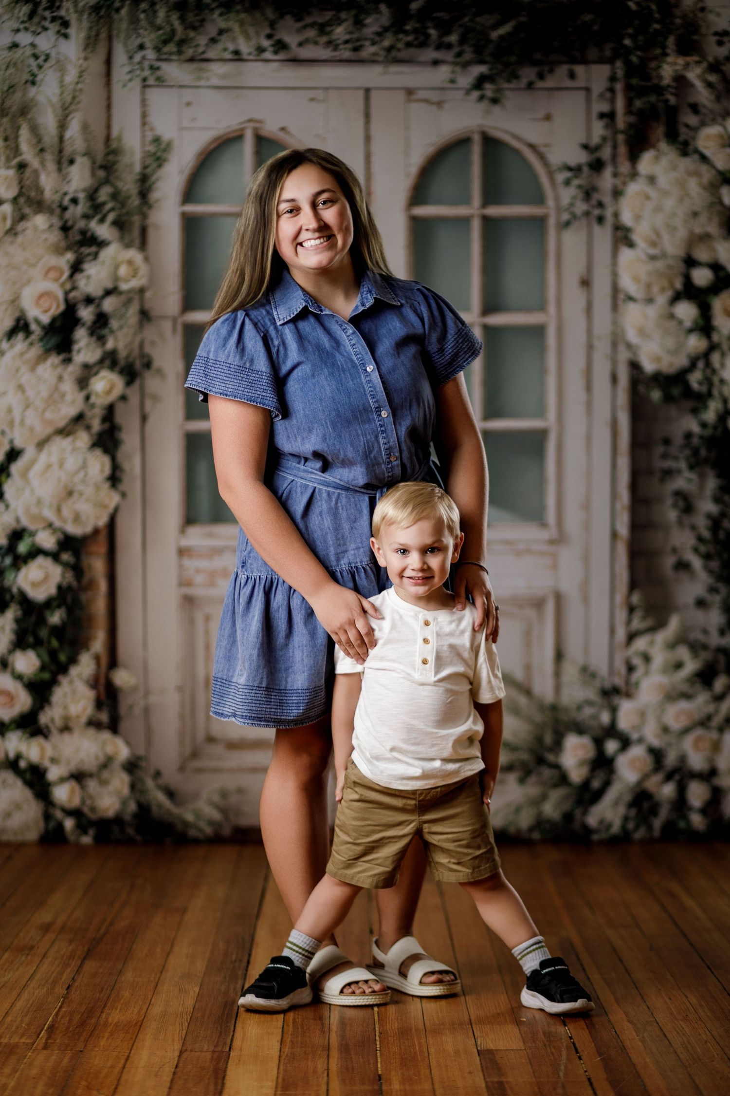 A smiling woman in a blue denim dress poses with a toddler boy in white shirt and khaki shorts before a floral backdrop.