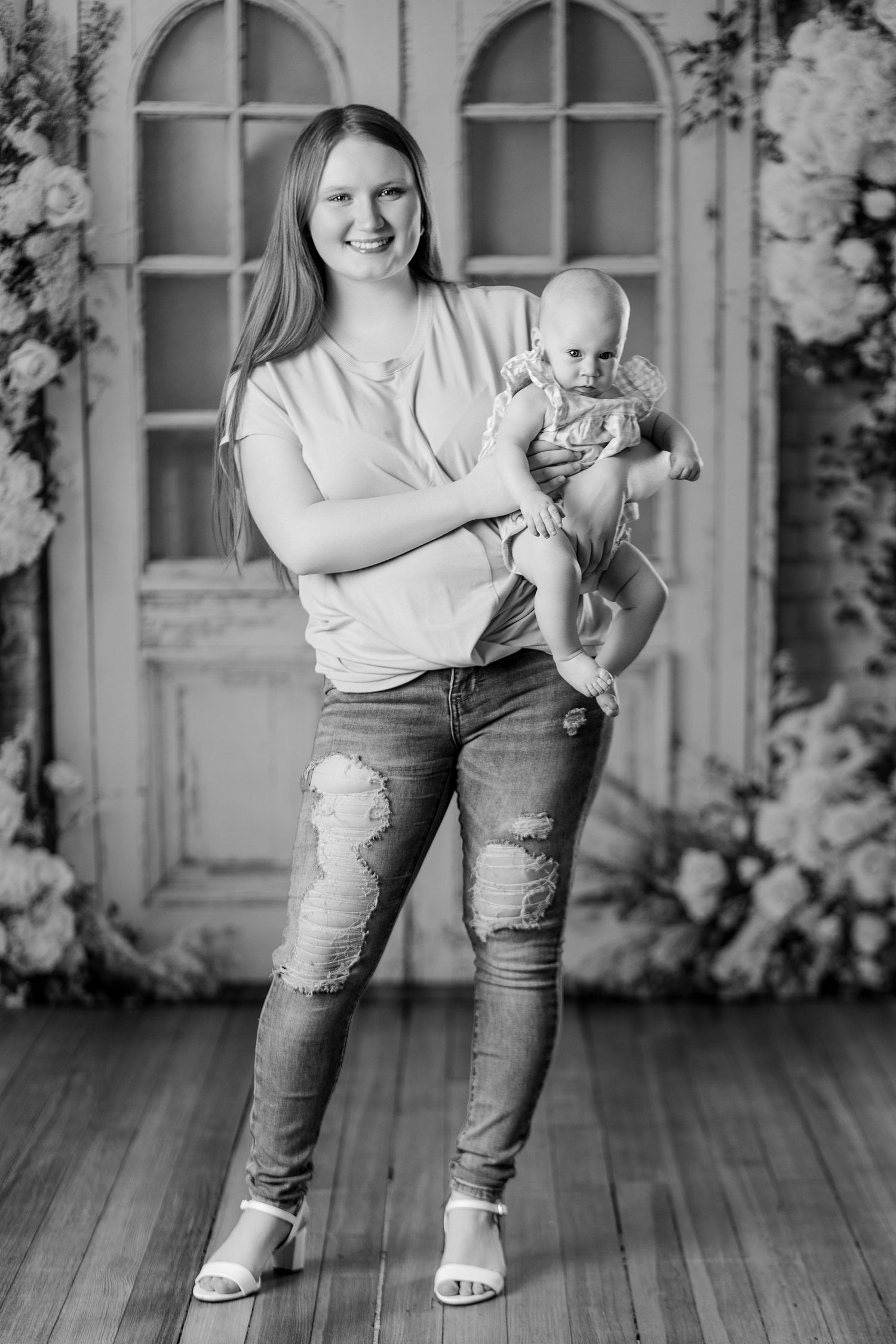Black and white portrait of a smiling mother holding her baby in a floral photography studio.