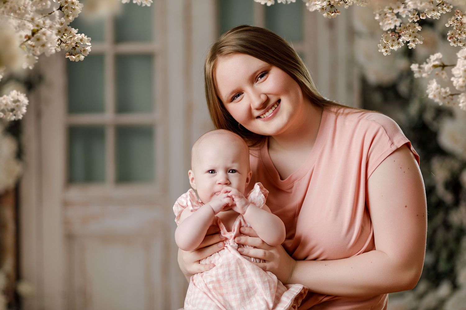 Smiling mother in pink top holds baby girl in floral spring photography studio setting.