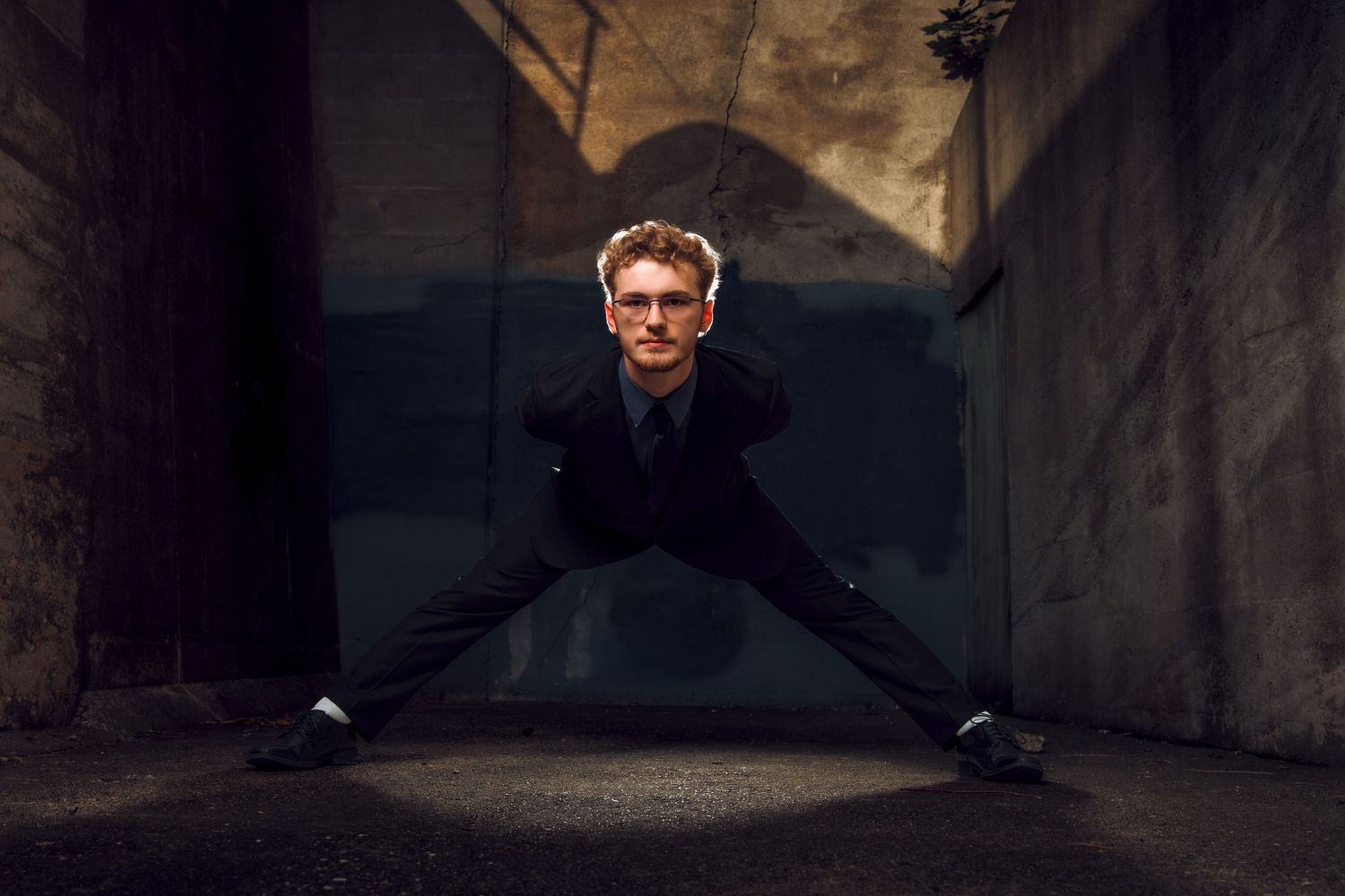 Young man in black suit doing wide leg split pose in dramatic concrete tunnel with moody lighting.