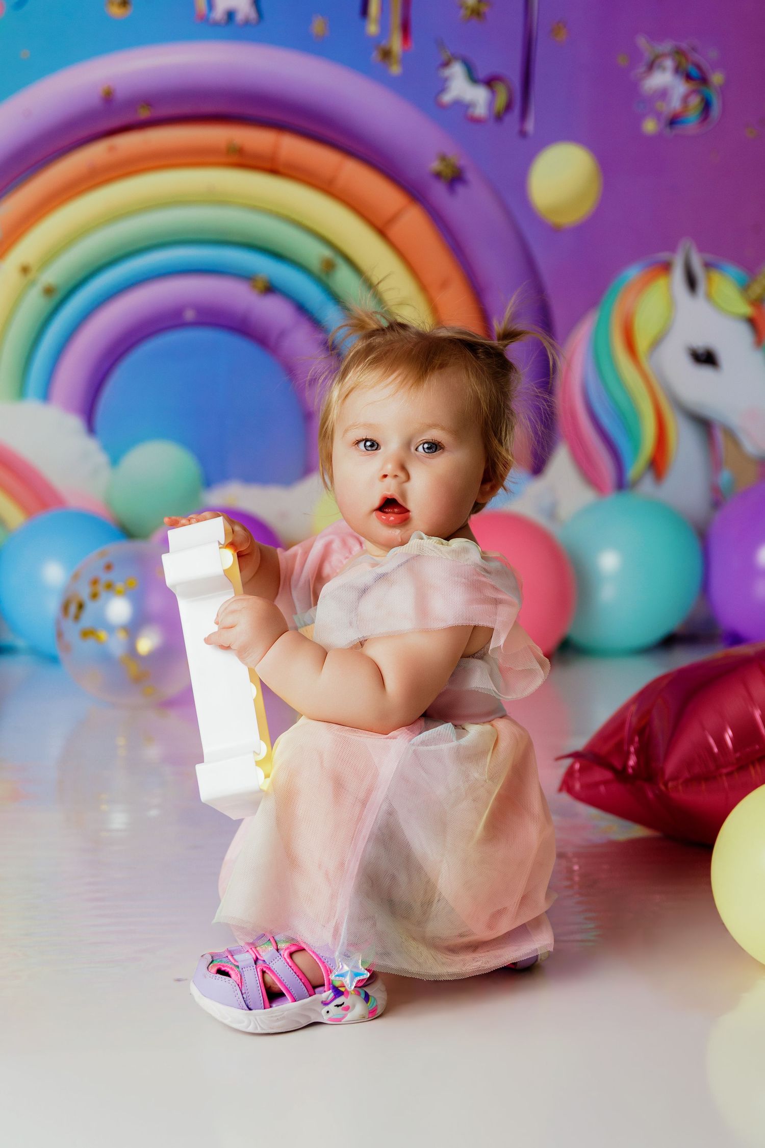 Toddler girl in pink dress holds cake tier at unicorn rainbow birthday party.