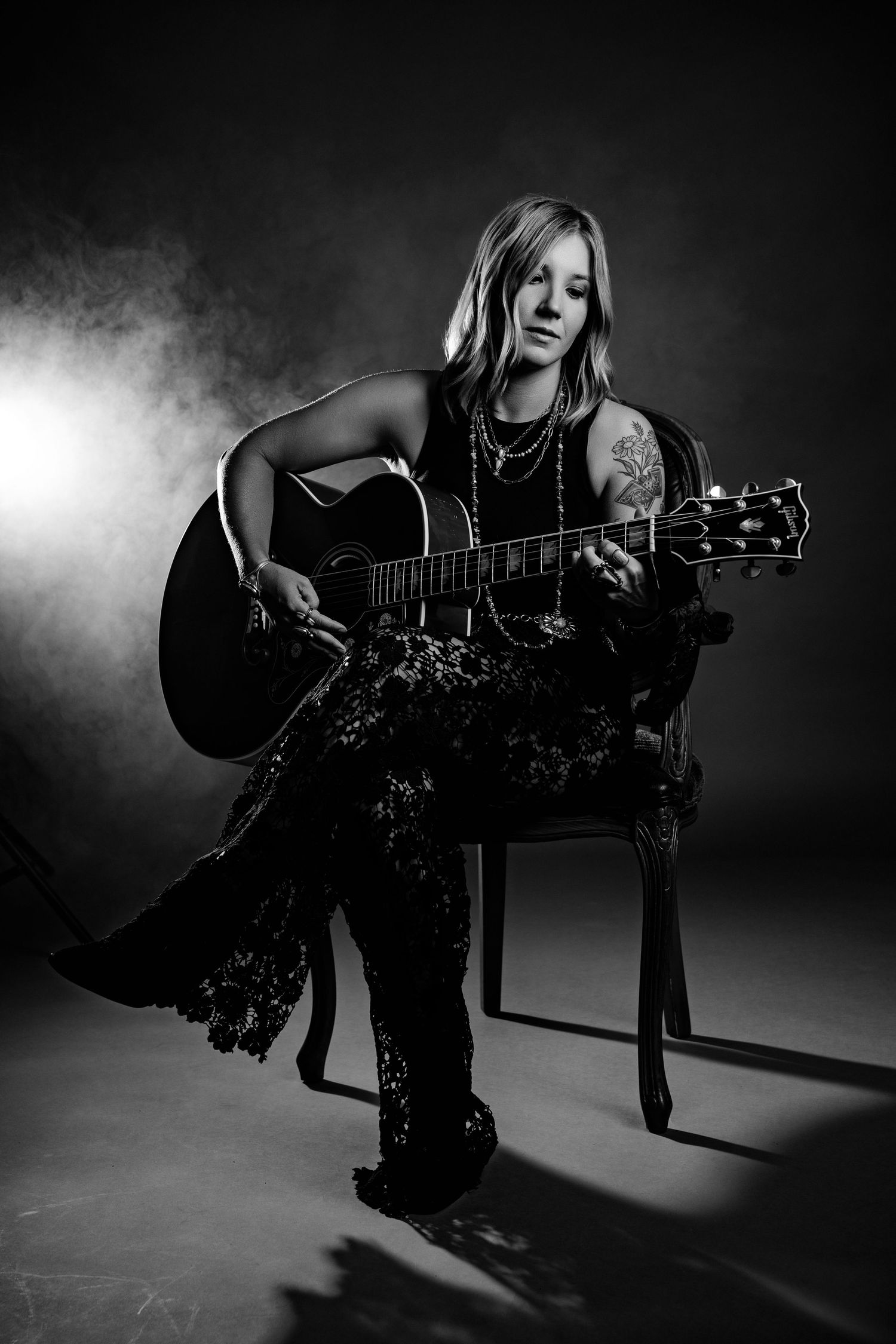 Black and white studio portrait of tattooed female musician playing acoustic guitar while seated.