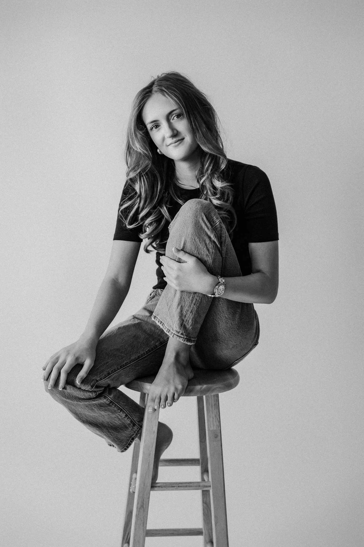 Smiling young woman in black t-shirt and jeans seated on stool, casual black and white studio photo.