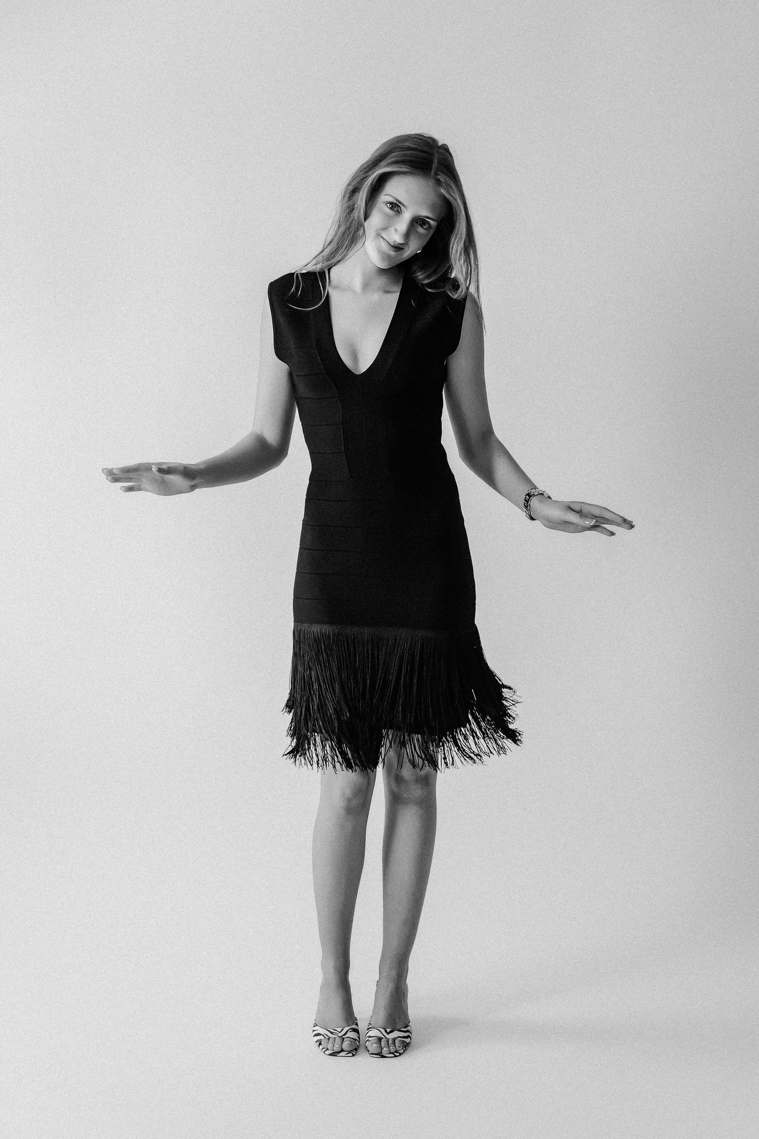 Woman in black fringe dress with v-neck poses playfully with arms outstretched in black and white photo.