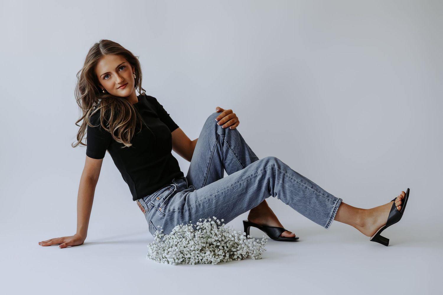 Woman in black tee and jeans sitting on floor beside white flowers wearing heels.