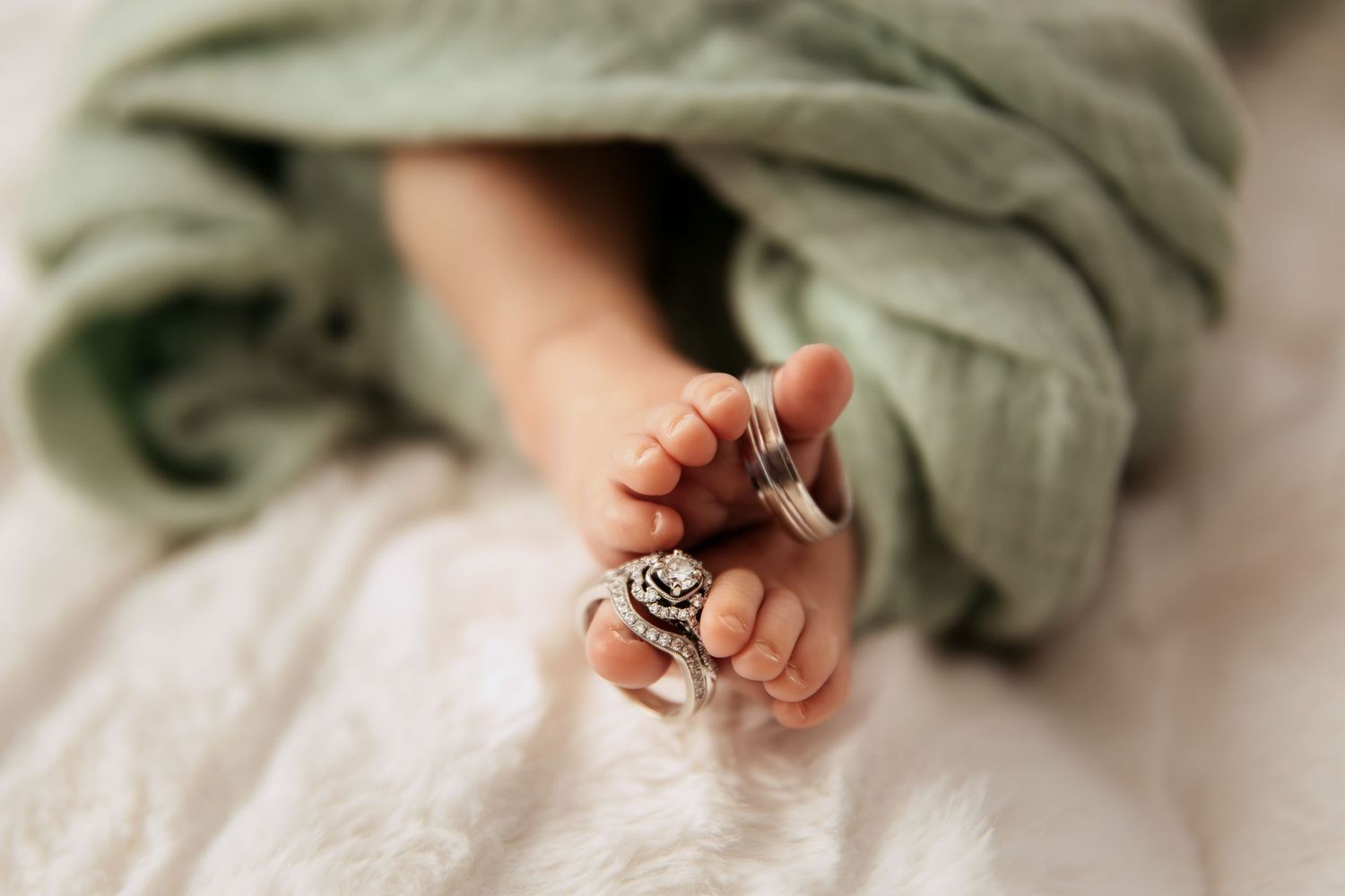 A tiny baby foot with a silver ring around the toe peeks out from under a sage green blanket.