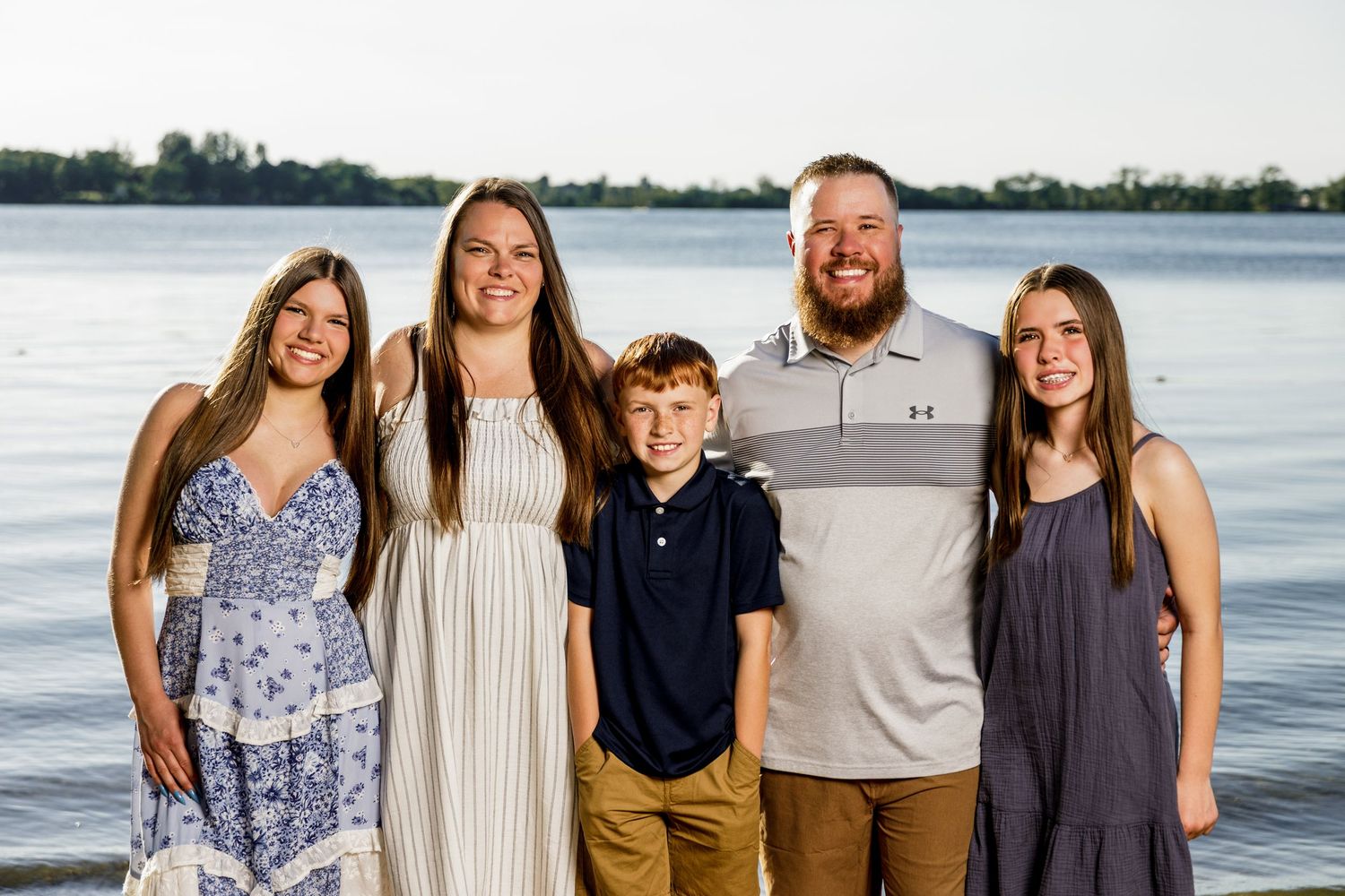 Family of five smiling together outdoors near a calm lake at sunset in casual summer attire.