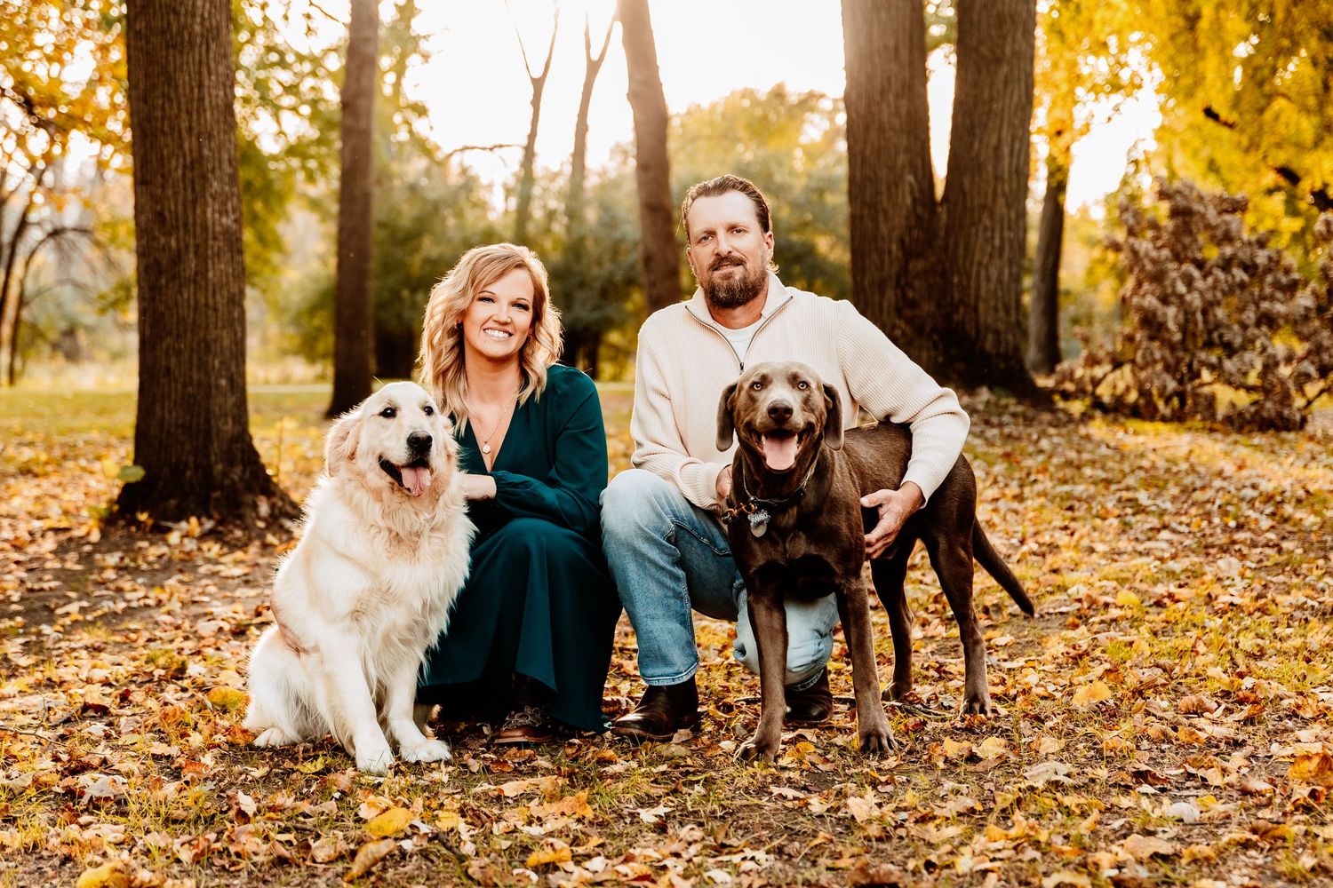 A family and their two dogs pose together on fallen autumn leaves in a wooded park setting during golden hour.